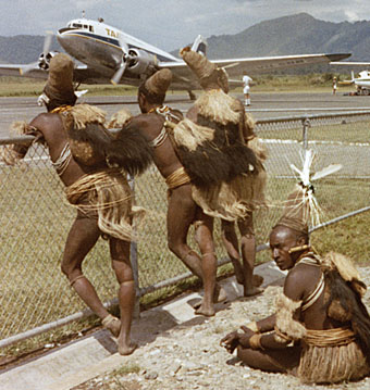 [3 Hewa men wearing ass grass, cassowary decorated feather bilum bags and domed cone-shaped hats lean on the chainlink fence watching the planes. Another sits cross-legged looking away.: 106k]